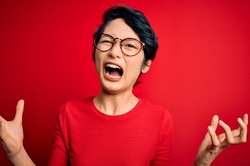 Young beautiful asian girl wearing casual t-shirt and glasses over isolated red background crazy and mad shouting and yelling with aggressive expression and arms raised. Frustration concept.