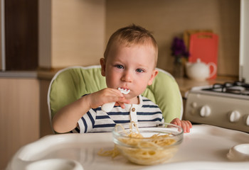 A little boy in a striped t-shirt sits in a high chair and eats pasta. Look at the camera.