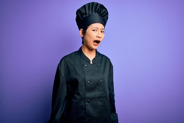 Young beautiful chinese chef woman wearing cooker uniform and hat over purple background In shock face, looking skeptical and sarcastic, surprised with open mouth