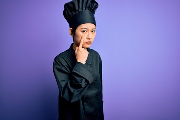 Young beautiful chinese chef woman wearing cooker uniform and hat over purple background Pointing to the eye watching you gesture, suspicious expression