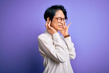 Young beautiful asian girl wearing casual shirt and glasses standing over purple background covering ears with fingers with annoyed expression for the noise of loud music. Deaf concept.