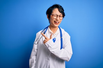 Fototapeta premium Young beautiful asian doctor girl wearing stethoscope and coat with blue cancer ribbon cheerful with a smile of face pointing with hand and finger up to the side with happy and natural expression
