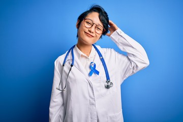 Young beautiful asian doctor girl wearing stethoscope and coat with blue cancer ribbon confuse and wonder about question. Uncertain with doubt, thinking with hand on head. Pensive concept.