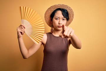 Young beautiful asian tourist girl on vacation wearing summer hat using hand fan pointing with finger to the camera and to you, hand sign, positive and confident gesture from the front