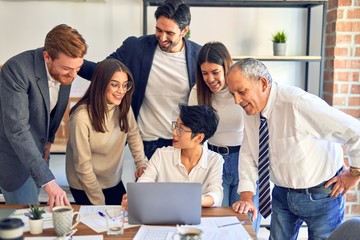 Group of business workers smiling happy and confident. One of them sitting and partners standing around. Working together with smile on face looking at the laptop at the office