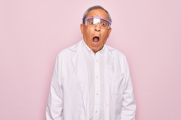 Senior grey-haired scientist man wearing glasses and coat over isolated pink background afraid and shocked with surprise expression, fear and excited face.