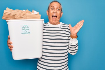 Senior man recycling holding trash can with cardboard to recycle over blue background very happy and excited, winner expression celebrating victory screaming with big smile and raised hands