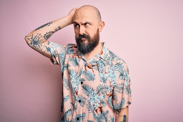 Handsome bald man with beard and tattoo wearing casual floral shirt over pink background confuse and wondering about question. Uncertain with doubt, thinking with hand on head. Pensive concept.