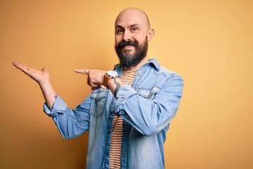 Handsome bald man with beard wearing casual denim jacket and striped t-shirt amazed and smiling to the camera while presenting with hand and pointing with finger.