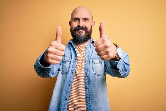 Handsome bald man with beard wearing casual denim jacket and striped t-shirt approving doing positive gesture with hand, thumbs up smiling and happy for success. Winner gesture.