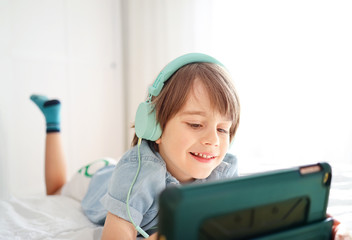 Cute little boy in casual clothes and headphones using a digital tablet and listening to music while sitting on bed at home