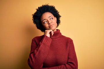 Young beautiful African American afro woman with curly hair wearing casual turtleneck sweater with hand on chin thinking about question, pensive expression. Smiling and thoughtful face. Doubt concept.