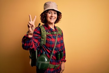 Fototapeta na wymiar Middle age curly hair hiker woman hiking wearing backpack and water canteen using binoculars smiling looking to the camera showing fingers doing victory sign. Number two.