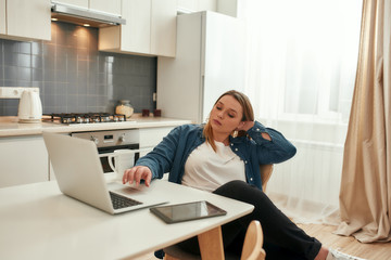 Pleasure of working from home. Young caucasian woman in casual clothes sitting at kitchen table at home and working on laptop. Working remotely