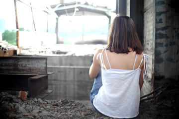 young woman in a white dress,  A girl brown hair in a white sleeveless T-shirt sits on the floor in an abandoned and ruined building, shot from behind
