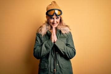 Middle age beautiful blonde skier woman wearing snow sportwear and ski goggles praying with hands together asking for forgiveness smiling confident.