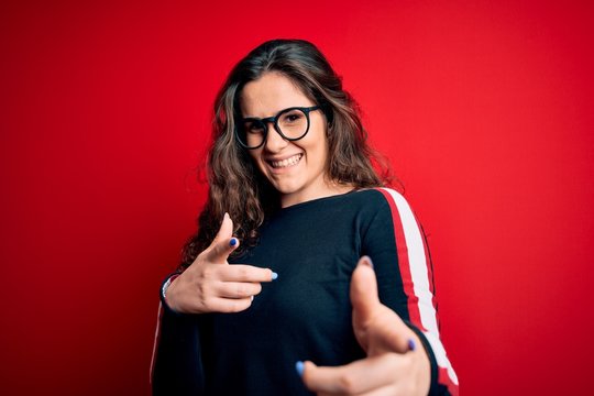 Young beautiful woman with curly hair wearing sweater and glasses over red background pointing fingers to camera with happy and funny face. Good energy and vibes.