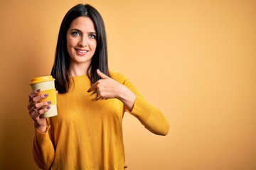 Young woman with blue eyes holding cup of coffee standing over yellow background with surprise face pointing finger to himself