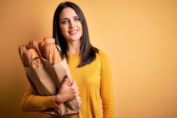 Young woman with blue eyes holding paper bag with bread over isolated yellow background with a happy face standing and smiling with a confident smile showing teeth