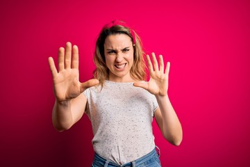 Young beautiful blonde woman wearing casual t-shirt standing over isolated pink background afraid and terrified with fear expression stop gesture with hands, shouting in shock. Panic concept.