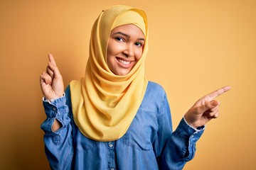 Young beautiful woman with curly hair wearing arab traditional hijab over yellow background smiling confident pointing with fingers to different directions. Copy space for advertisement