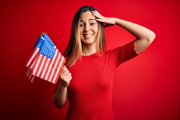 Young beautiful blonde patriotic woman with blue eyes holding united states flags stressed with hand on head, shocked with shame and surprise face, angry and frustrated. Fear and upset for mistake.