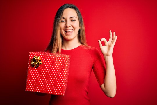 Young beautiful blonde woman with blue eyes holding birthday gift over red background doing ok sign with fingers, excellent symbol