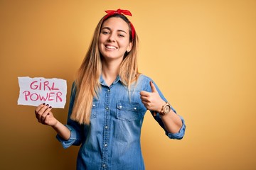 Blonde woman with blue eyes asking for girls rights holding banner with girl power message happy with big smile doing ok sign, thumb up with fingers, excellent sign