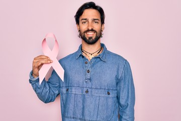 Young handsome hispanic man holding breast cancer awareness pink ribbon with a happy face standing...