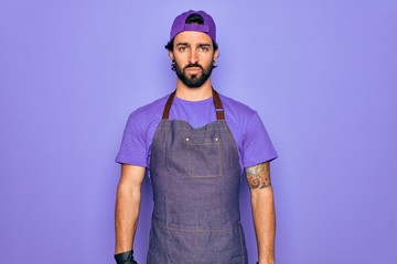 Young handsome hispanic man with bear wearing professional apron working as tattoo artist with serious expression on face. Simple and natural looking at the camera.