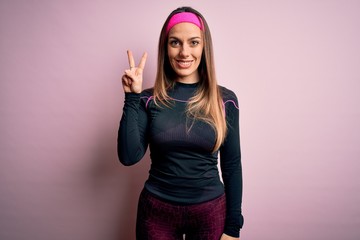 Young blonde fitness woman wearing sport workout clothes over isolated background smiling looking to the camera showing fingers doing victory sign. Number two.