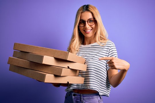 Young beautiful blonde woman holding boxes of italian pizza over isolated purple background very happy pointing with hand and finger