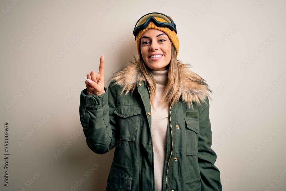 Poster young brunette skier woman wearing snow clothes and ski goggles over white background showing and po