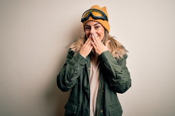 Young brunette skier woman wearing snow clothes and ski goggles over white background laughing and embarrassed giggle covering mouth with hands, gossip and scandal concept