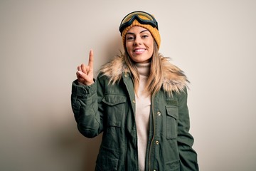 Young brunette skier woman wearing snow clothes and ski goggles over white background showing and pointing up with finger number one while smiling confident and happy.