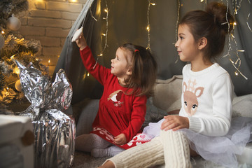 Little sisters unpack gifts smile laugh Cozy atmosphere of Christmas New Year Christmas tree garlands and a fireplace Dreams come true Children Baby