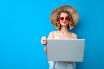 girl in a sun hat and glasses use a laptop on a blue isolated background, a woman with a computer...