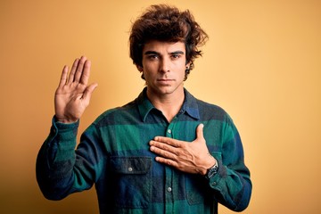 Young handsome man wearing casual shirt standing over isolated yellow background Swearing with hand...