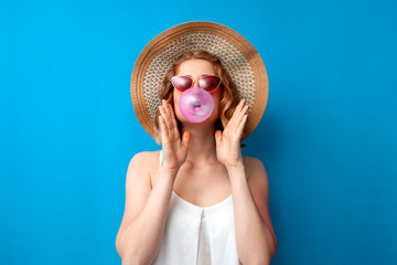 girl in a sun hat and glasses inflates a bubble gum on a blue isolated background, a woman chews a...