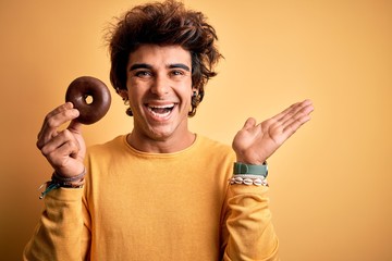 Young handsome man holding chocolate donut standing over isolated yellow background very happy and...