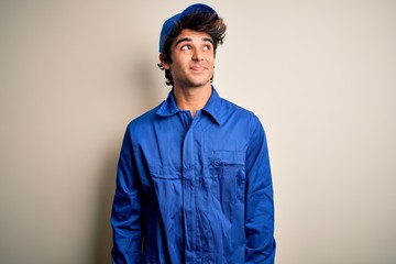 Young mechanic man wearing blue cap and uniform standing over isolated white background smiling looking to the side and staring away thinking.