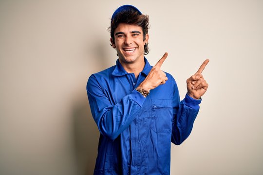 Young mechanic man wearing blue cap and uniform standing over isolated white background smiling and looking at the camera pointing with two hands and fingers to the side.