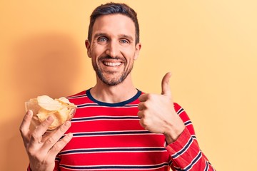 Young handsome man holding bowl of potato chips standing over isolated yellow background smiling happy and positive, thumb up doing excellent and approval sign