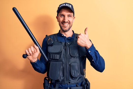 Handsome policeman wearing uniform and bulletprof holding baton over yellow background smiling happy and positive, thumb up doing excellent and approval sign