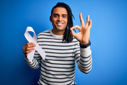 Young african american man with dreadlocks holding white ribbon over blue background doing ok sign with fingers, excellent symbol