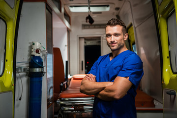 Handsome paramedic in a medical uniform looking at the camera and smiling, ambulance car in the background