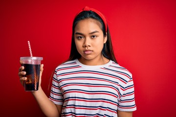 Young asian girl drinking cola fizzy refreshment using straw over isolated red background with a confident expression on smart face thinking serious