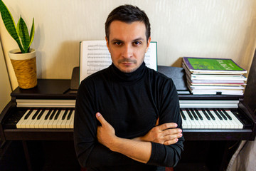 A dark-haired tanned young guy wearing a black turtleneck sitting with arms crossed with his back to the digital piano, looking directly into the camera.