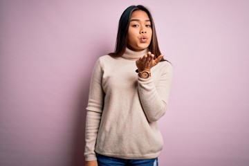 Young beautiful asian girl wearing casual turtleneck sweater over isolated pink background looking at the camera blowing a kiss with hand on air being lovely and sexy. Love expression.