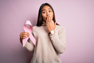 Young asian girl holding pink cancer ribbon symbol for suppport over isolated background cover mouth with hand shocked with shame for mistake, expression of fear, scared in silence, secret concept